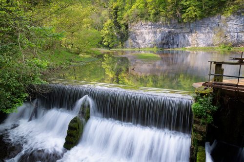 Long exposure of Cressbrook weir in full flow with fresh green spring vegetation.